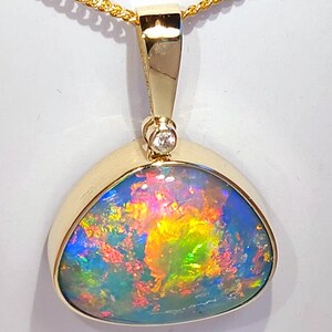 Puede incluir: Colgante de oro con una piedra preciosa de ópalo. El ópalo muestra un espectro de colores, incluyendo azul, verde, amarillo y rojo. Un pequeño diamante está engastado encima del ópalo. El colgante cuelga de una cadena de oro.