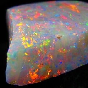 Könnte beinhalten: Nahaufnahme eines rohen Opal-Edelsteins. Der Opal zeigt eine milchig-weiße Grundfarbe mit lebendigen Blitzen in Orange, Gelb, Grün, Blau und Rosa. Der Edelstein hat eine raue, natürliche Textur und eine unregelmäßige Form.