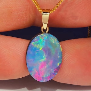 May include: A gold pendant necklace with an oval opal gemstone. The opal showcases a vibrant play of colour, with shades of blue, pink, and green. The chain is a delicate gold link. The pendant is held between two fingers.