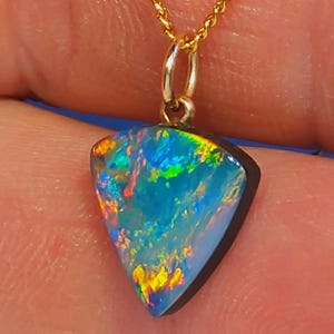 May include: A pendant necklace with a triangular opal gemstone, displaying blue, green, and yellow colours. The opal is set in a dark bezel and hangs from a delicate gold chain. The gemstone exhibits a play of colour.