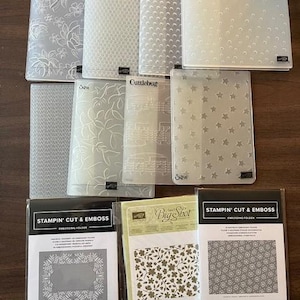 May include: A collection of clear embossing folders with various patterns, including floral, dotted, and star designs. Several folders are packaged with the "Stampin' Cut & Emboss" and "Big Shot" labels. The folders are arranged on a wooden surface.