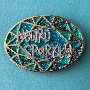 Op de afbeelding: Een ovale speld met een gouden rand en de woorden "NEURO SPARKLY" in zilveren letters. Het midden heeft een sprankelend turquoise en blauw ontwerp, met geometrische vormen. De speld staat tegen een turquoise achtergrond.