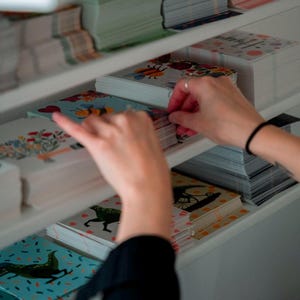 May include: A white shelf unit filled with stacks of colorful greeting cards. The cards feature various illustrations, including bees, flowers, and animals. A person's hand is reaching for a stack of cards.