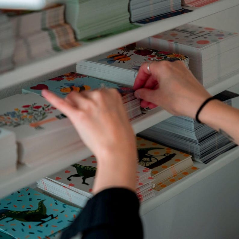 May include: A white shelf unit filled with stacks of colorful greeting cards. The cards feature various illustrations, including bees, flowers, and animals. A person's hand is reaching for a stack of cards.