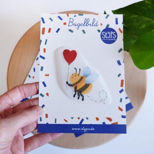 Può includere: Una carta bianca con un bordo di coriandoli colorati presenta un'ape dei cartoni animati che tiene in mano un palloncino a forma di cuore rosso. L'ape è gialla e nera con ali blu. La carta ha il testo "Bügelbild" e "STYPS".