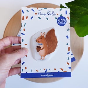 Könnte beinhalten: Eine weiße Karte mit einem Cartoon-Eichhörnchen, das eine Eichel hält und eine blaue Fliege trägt. Die Karte trägt den Text "Bügelbild" und "styps" mit einer Website-Adresse unten. Die Karte ist mit blauen und orangefarbenen Konfetti verziert.