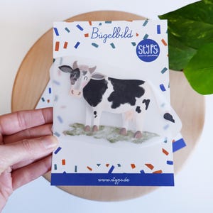 Könnte beinhalten: Eine Packung mit einem Bügelbild einer Cartoon-Kuh. Die Kuh ist schwarz und weiß mit einer rosa Nase und steht auf grünem Gras. Die Packung trägt den Text "Bügelbild" und "www.atypo.de".