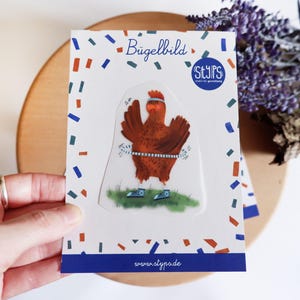 Può includere: Un adesivo con un pollo dei cartoni animati con una fascia e scarpe da ginnastica, che fa esercizio. L'adesivo è su una carta bianca con coriandoli e il testo "Bügelbild" e "STYPS". È visibile anche il sito web www.styps.de.