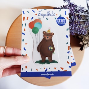 Puede incluir: Una tarjeta de felicitación con un oso pardo con gafas y un gorro de fiesta, sosteniendo globos y flores. La tarjeta tiene confeti y el texto "Bügelbild" y "STYPS". La tarjeta está sujeta por una mano.