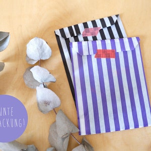 May include: Two striped paper gift bags with red and white tape. The bags are black and white striped and purple and white striped. The text "BUNTE VERPACKUNG!" is on a purple circle in the lower left corner of the image.