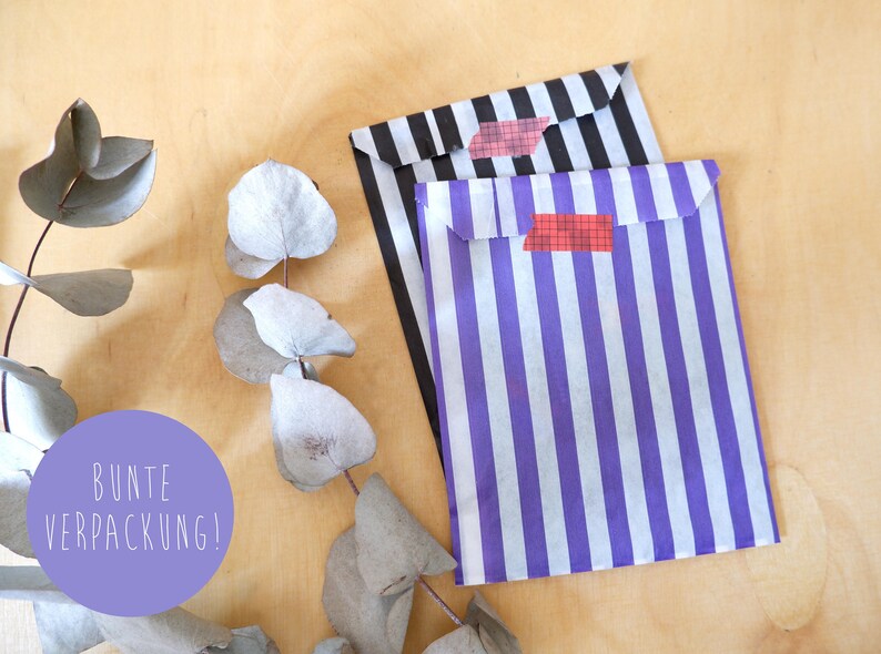 May include: Two striped paper gift bags with red and white tape. The bags are black and white striped and purple and white striped. The text "BUNTE VERPACKUNG!" is on a purple circle in the lower left corner of the image.