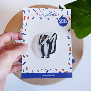 Può includere: Una carta bianca con un disegno di un tasso con gli occhiali. La carta ha un bordo blu con coriandoli e il testo "Bügelbild" e "STYPS" in un cerchio. Il sito web www.styps.de è stampato in basso.