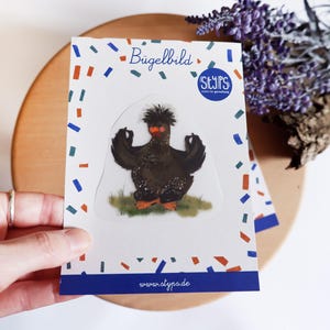 Op de afbeelding: Een sticker met een zwarte kip met een rood gezicht en oranje poten, in een meditatieve pose. De sticker staat op een witte achtergrond met het woord "Bügelbild" en kleurrijke confetti. De website www.atypo.de is onderaan zichtbaar.