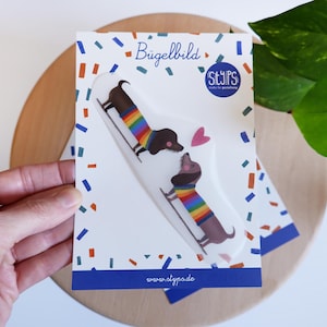 Könnte beinhalten: Eine weiße Karte mit zwei Bügelbildern von Dackel-Hunden. Die Hunde sind braun mit Regenbogen-gestreiften Pullovern. Die Karte trägt den Text "Bügelbild" und "styps" in einem blauen Kreis, mit Konfetti und einem blauen Rand.