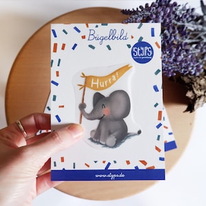 Könnte beinhalten: Ein Aufkleber mit einem grauen Cartoon-Elefanten, der ein Banner mit der Aufschrift "Hurra!" hält. Der Aufkleber befindet sich auf einem weißen Hintergrund mit Konfetti und einem blauen Rand. Der Markenname "STYPS" ist sichtbar.