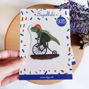 Könnte beinhalten: Weiße Karte mit einem grünen Dinosaurier, der einen orangefarbenen Helm trägt und Fahrrad fährt. Die Karte trägt den Text "Bügelbild" und ein "STYPS"-Logo. Der Hintergrund ist mit Konfetti und der Website-Adresse www.styps.de verziert.