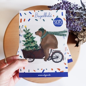 Könnte beinhalten: Eine transparente Kunststoffverpackung mit einer skurrilen Illustration eines braunen Bären, der Fahrrad fährt und einen Wagen mit einem Weihnachtsbaum zieht. Der Bär trägt eine grüne Mütze und einen Schal. Der Text "Bügelbild" steht oben.