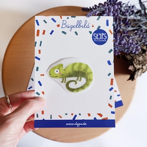 May include: A green chameleon iron-on patch on a white card with the text "Bügelbild" and "styps". The card has a blue border and colorful confetti-like accents. The website www.styps.de is also visible.