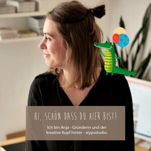 May include: A woman with brown hair looks to the side. She is wearing a black shirt. A green alligator with red and blue balloons is on her shoulder. The text "HI, SCHON DASS DU HIER BIST! Ich bin Anja - Gr&uuml;nderin und der kreative Kopf hinter - stypsstudio." is below her.