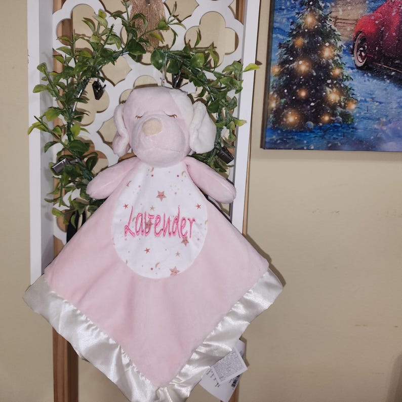 May include: A pink plush dog-shaped baby security blanket with the name "Lavender" embroidered on the front. The blanket has a satin border and a white bib-like section with star and moon designs.