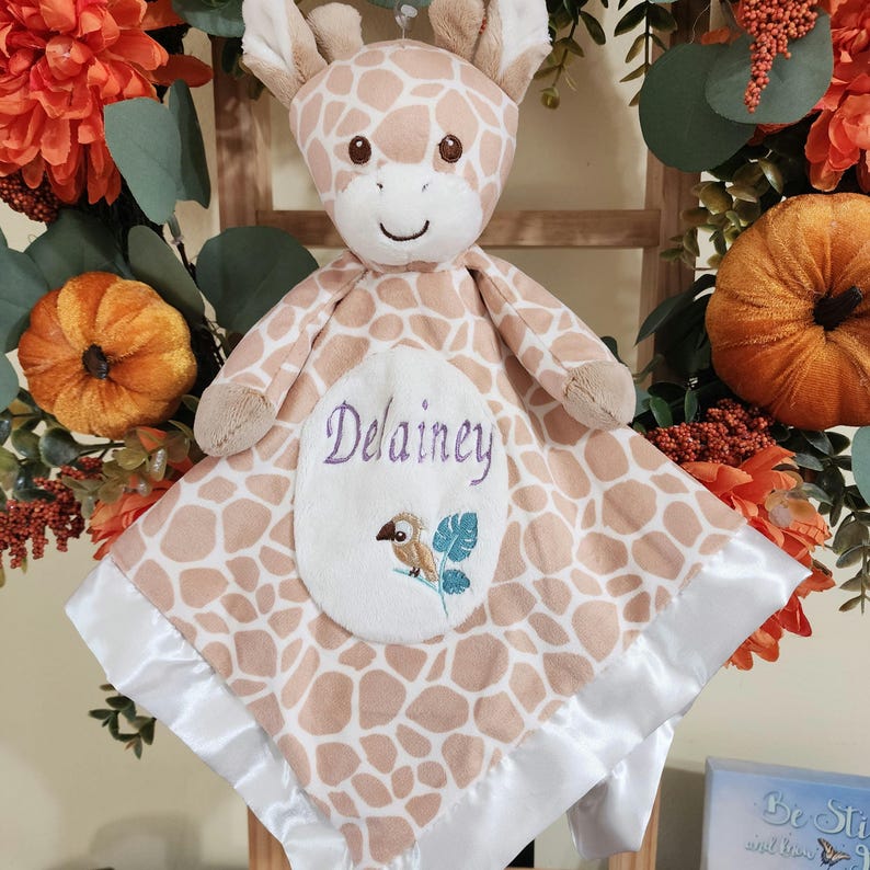 May include: A plush giraffe-shaped baby blanket with a giraffe pattern in shades of tan and cream. The blanket features a white satin trim and an embroidered patch with the name "Delainey" and a bird design.