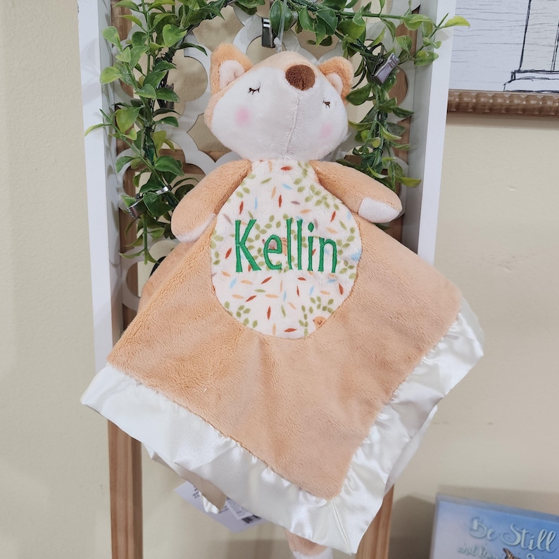 May include: A plush fox-shaped baby security blanket in peach and white. The blanket features a satin trim and a patterned patch with the name "Kellin" embroidered in green. The fox has closed eyes and a brown nose.
