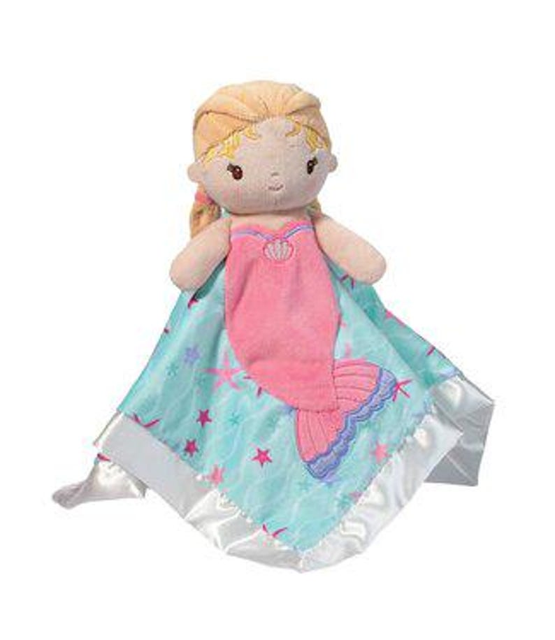 May include: A plush mermaid doll with blonde hair and a pink tail, wearing a pink seashell top, resting on a light blue blanket with a star and wave pattern. The blanket has a satin border.