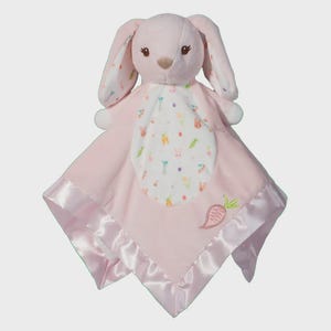 May include: A pink plush bunny security blanket. The bunny has long ears with a floral pattern, and a soft pink blanket with a satin trim. The blanket features a white center with bunny and carrot designs.