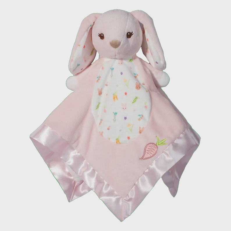 May include: A pink plush bunny security blanket. The bunny has long ears with a floral pattern, and a soft pink blanket with a satin trim. The blanket features a white center with bunny and carrot designs.