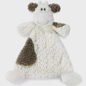 May include: A white and brown plush cow-shaped baby blanket. The blanket features a textured, fluffy white body with brown patches and accents. The cow has a smiling face with blue eyes and brown ears.