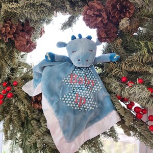 May include: A blue plush baby blanket with a dragon head and arms. The blanket has a white satin border and a patterned center with the words "Baby Ip" in pink. The blanket is displayed against a backdrop of a Christmas wreath with pinecones and red berries.