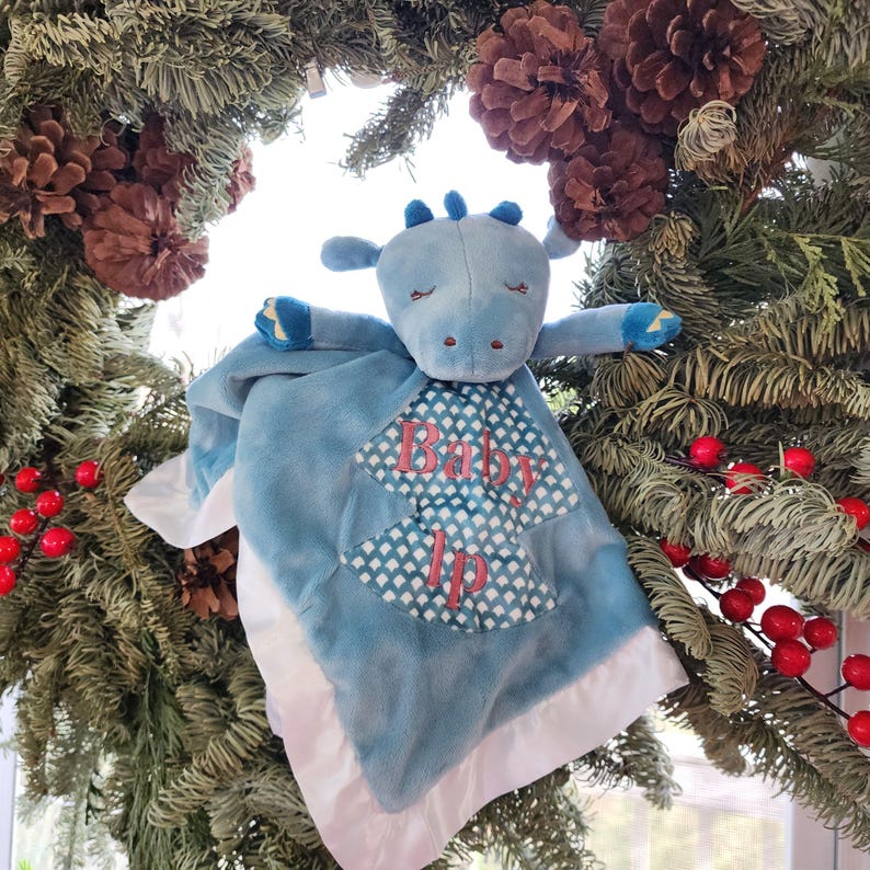 May include: A blue plush baby blanket with a dragon head and arms. The blanket has a white satin border and a patterned center with the words "Baby Ip" in pink. The blanket is displayed against a backdrop of a Christmas wreath with pinecones and red berries.