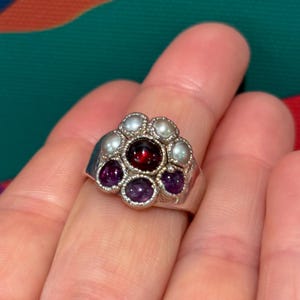 May include: A silver ring featuring a floral design. The ring's center stone is a deep red, surrounded by six smaller purple stones and six white pearls. The ring is set against a colorful background.
