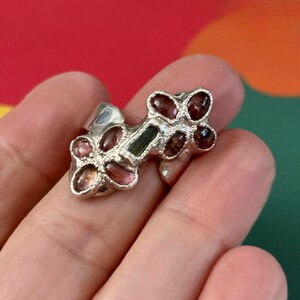 May include: A silver ring with a unique design featuring multiple pink and green gemstones. The ring is set on a finger.