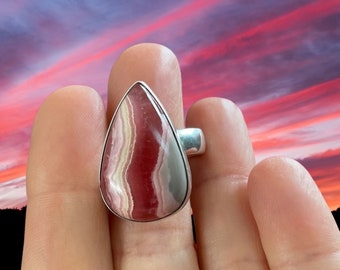 Puede incluir: Un anillo en forma de lágrima con una piedra preciosa de bandas rosadas y blancas engastada en una banda plateada.