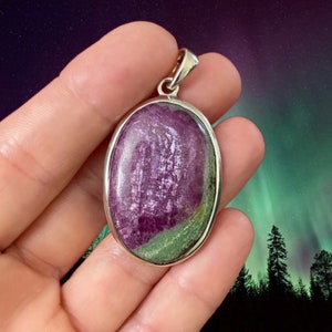 Puede incluir: Un colgante de plata con una piedra ovalada verde y morada. El colgante está engastado en un bisel de plata y tiene un aro para sujetar una cadena.