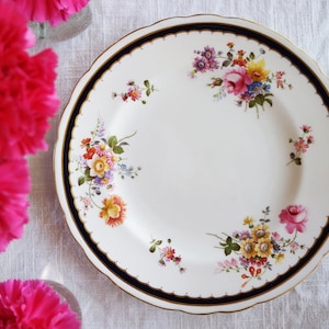 May include: A white porcelain dinner plate with a gold rim and a blue and gold band around the edge. The plate is decorated with a floral pattern in shades of pink, yellow, and green.