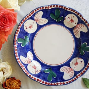 May include: A hand-painted ceramic plate with a blue border, white center, and fish and leaf designs. The plate features a scalloped edge with red dots. Decorative tableware.