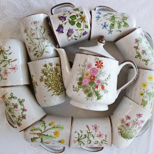 Puede incluir: Un juego de té de cerámica blanca con una tetera y tazas a juego. La tetera y las tazas están decoradas con diseños florales de varios colores. Las tazas tienen bordes y asas marrones. El juego tiene un acabado moteado.