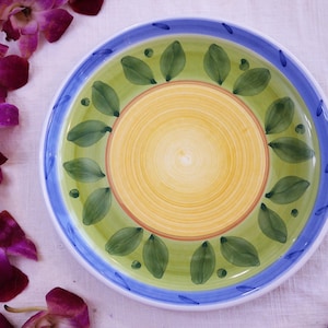 May include: A round ceramic plate with a yellow center and a green and blue border. The plate features a leaf design around the edge. The plate is decorated with a blue and green color scheme.