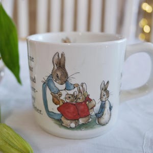 Könnte beinhalten: Weiße Keramiktasse mit einer Peter Rabbit-Illustration. Das Design zeigt Hasen in blauer und roter Kleidung. Die Tasse hat einen gebogenen Griff und ein gedrucktes Zitat.