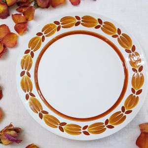 May include: White ceramic plate with a brown border and a repeating pattern of stylized orange and brown floral designs. The plate has a clean, minimalist aesthetic, suitable for serving food.