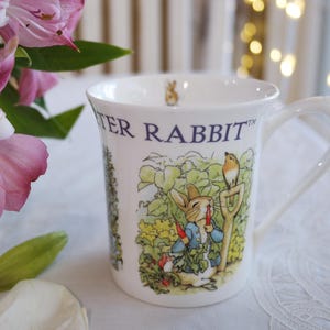 Könnte beinhalten: Ein weißer Keramikbecher mit einer farbigen Illustration von Peter Hase, der eine Karotte in einem Garten hält, während ein kleiner Vogel auf einer Schaufel sitzt. Der Text PETER RABBIT ist blau am oberen Rand gedruckt, innen ist ein kleines Häschen.