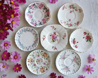 May include: A collection of seven white ceramic saucers arranged in a group. Each saucer features a unique floral pattern with various colors including pink, red, green, and blue. The saucers have scalloped or smooth edges with gold trim.