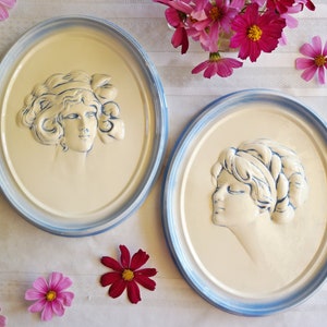May include: Two white ceramic wall plaques with blue trim, each featuring a raised relief of a woman's face. The plaques are on a white background with pink flowers.