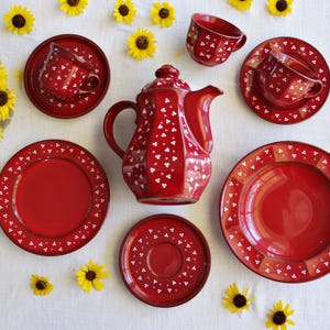 May include: A red ceramic tea set with white heart-shaped accents, including a teapot, cups, saucers, and plates. The set is arranged on a white surface, surrounded by yellow sunflowers.