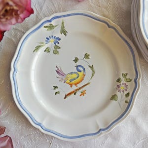 May include: A white ceramic plate with a blue rim and a hand-painted design of a yellow bird with blue accents and flowers. The plate is part of a set of plates.
