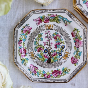 May include: A white octagonal porcelain plate with a gold rim and a floral design. The center of the plate features a stylized tree with pink and blue flowers. The border of the plate is decorated with a repeating pattern of pink flowers and green leaves.