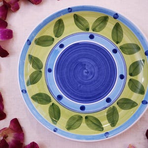 May include: A round, hand-painted ceramic plate with a vibrant design. The plate features a blue center with concentric circles, surrounded by a light green band with green leaf motifs. A blue and light blue border completes the design. The plate is approximately 10 inches in diameter.