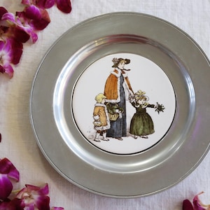 Puede incluir: Un plato de metal plateado con un inserto cerámico central. El inserto presenta una ilustración vintage de una mujer y dos niños. La mujer lleva sombrero y capa, tomados de la mano de los niños. Los niños llevan abrigos y vestidos.